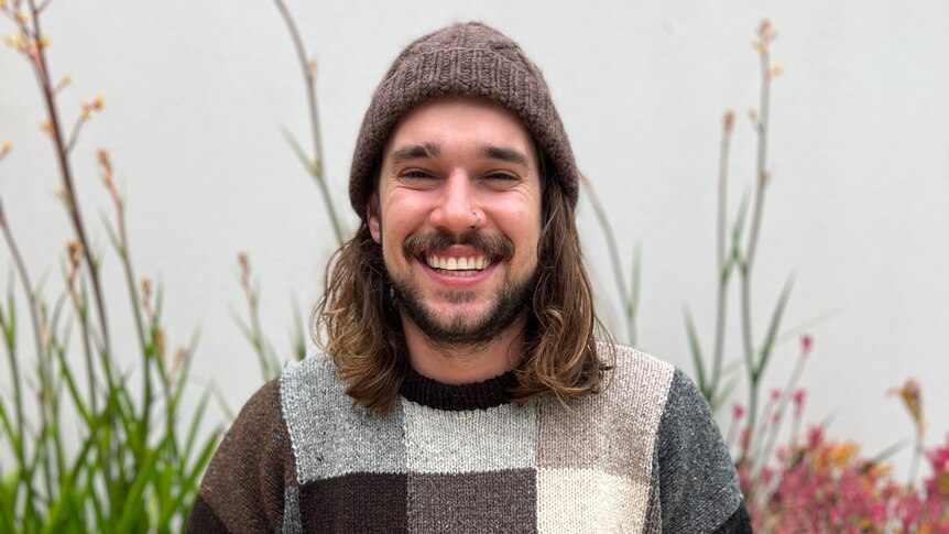 A man with long hair sits smiling at the camera. He is wearing a black and white checked jumper and a beanie. 
