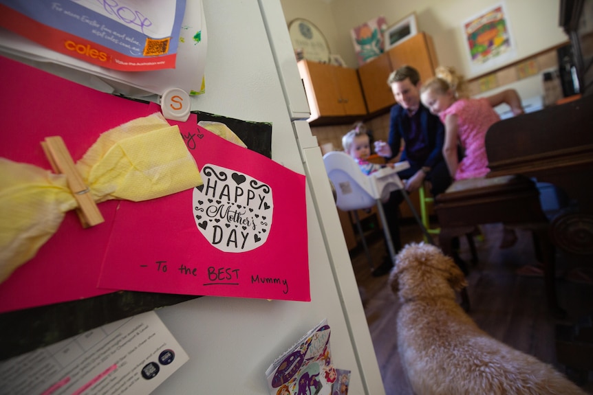 Bright pink hand made Mother's Day card on fridge, family in kitchen in the background