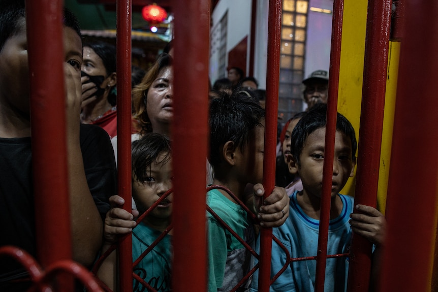 Kids and adults queue against vertical, red metal bars as they wait for food to break their fast.