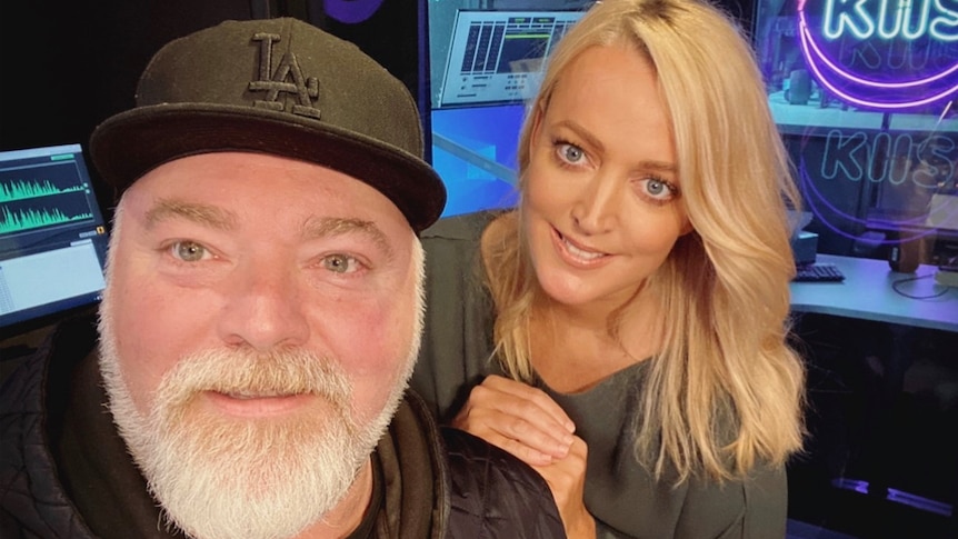 A man with a grey beard and black cap and a blonde woman resting her hand on his shoulder smile in a radio studio.