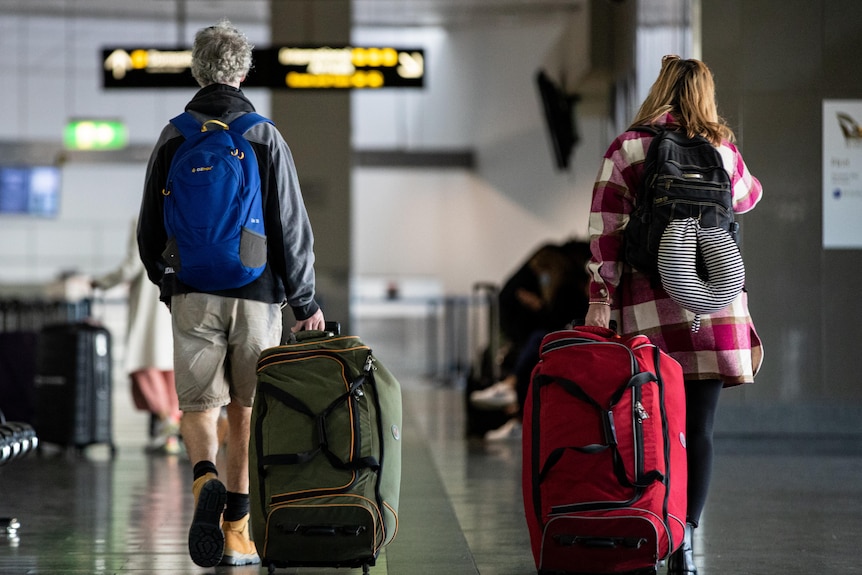 A man and a woman with backpacks and suitcases walk through an airport with their backs to the camera.