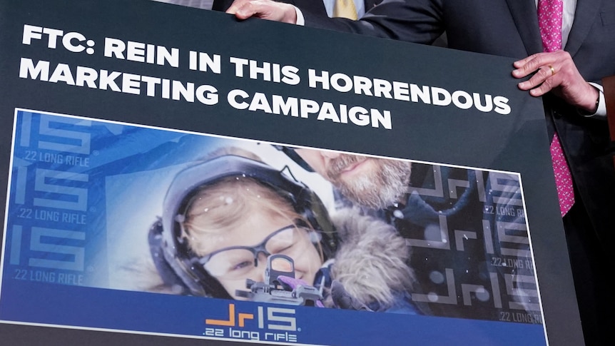 A man holds a sign reading 'FTC: Rein in this horrendous marketing campaign' with a child holding a gun