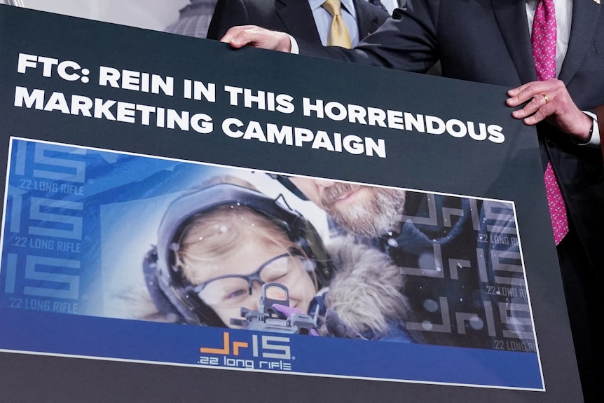 A man holds a sign reading 'FTC: Rein in this horrendous marketing campaign' with a child holding a gun