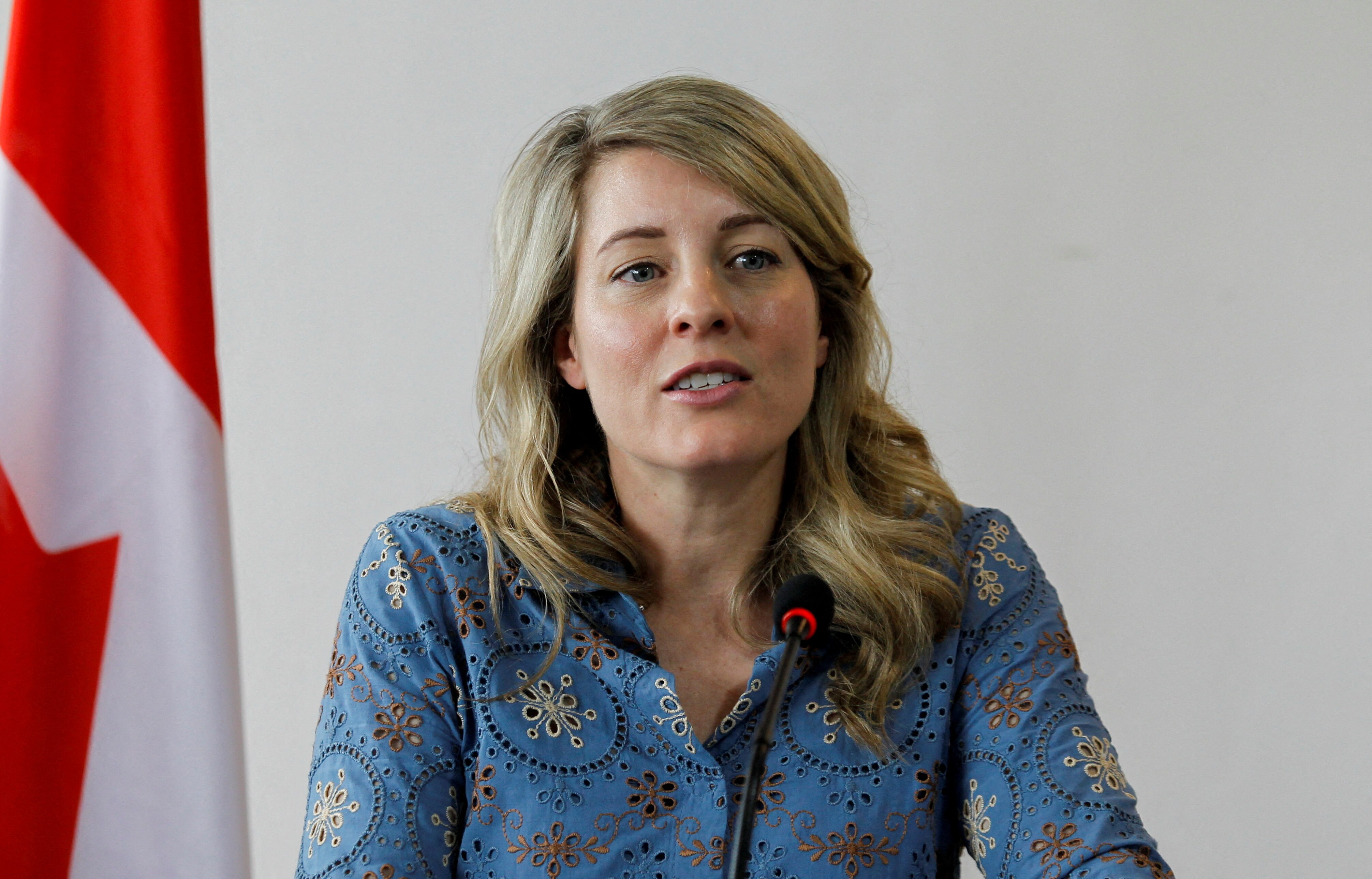 Foreign Minister, with blue shirt and blonde long hair, sits in front of microphone and Canadian flag.