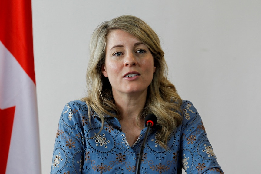 Foreign Minister, with blue shirt and blonde long hair, sits in front of microphone and Canadian flag.