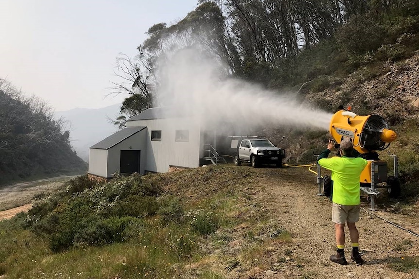 A yellow machine shoots out a cloud of water towards a building in mountainous bushland.