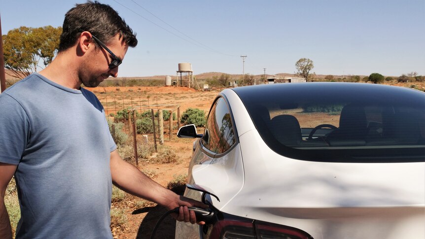 A man with short brown hair, wearing a blue T-shirt, charges an electric vehicle on an outback station.