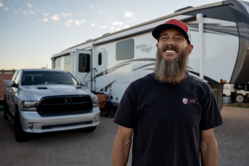 A man with a lush grey beard smiles widely, standing in front of a ute and large motor home.