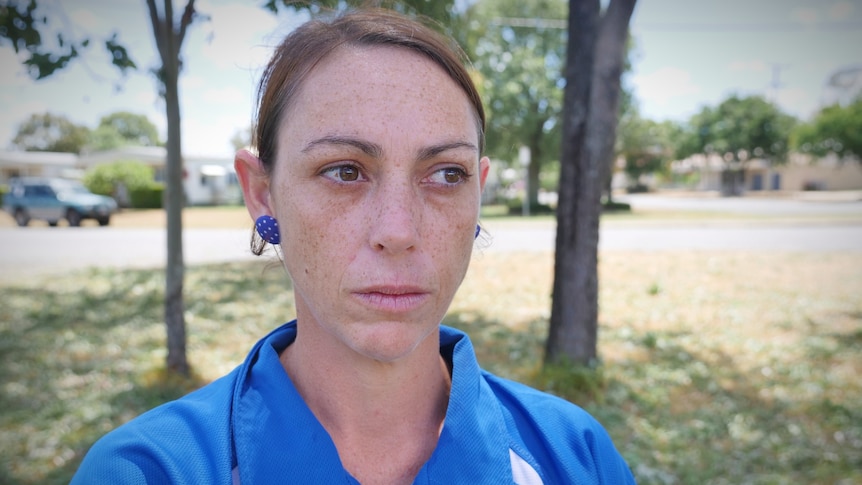 A woman in a blue collard shirt with brown hair looking just past the camera, standing outside. 