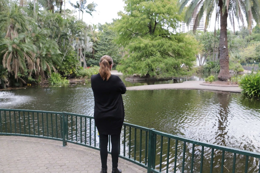 A woman stands with her arms folded looking over a pond in a park
