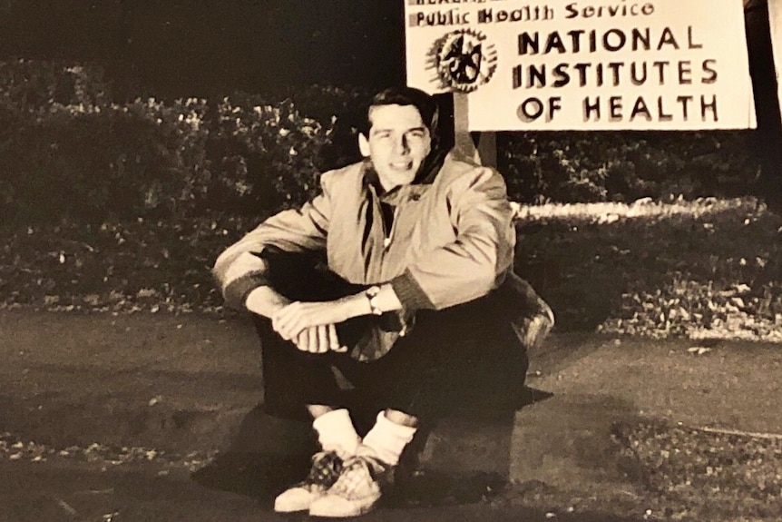 An old photograph of a young man sitting in front of a sign which reads "National Institutes of Health"