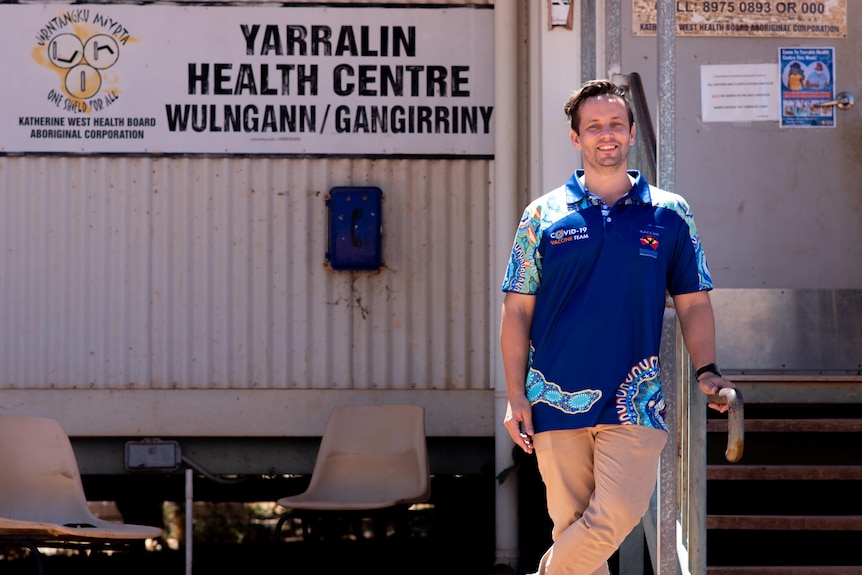 A smiling man in a blue shirt featuring an Aboriginal design, standing in front of a demountable building.