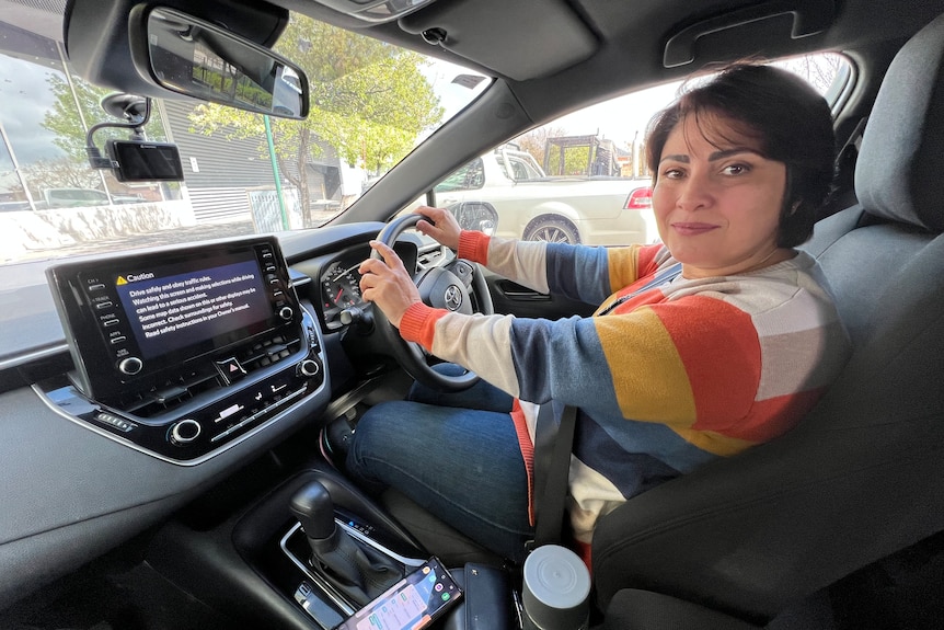 women with brown hair and rainbow top, friendly face, sitting in her car