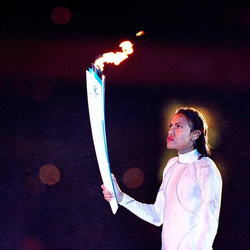 Australian athlete Cathy Freeman holds the lit Olympic torch up to the sky, with a dark background. 