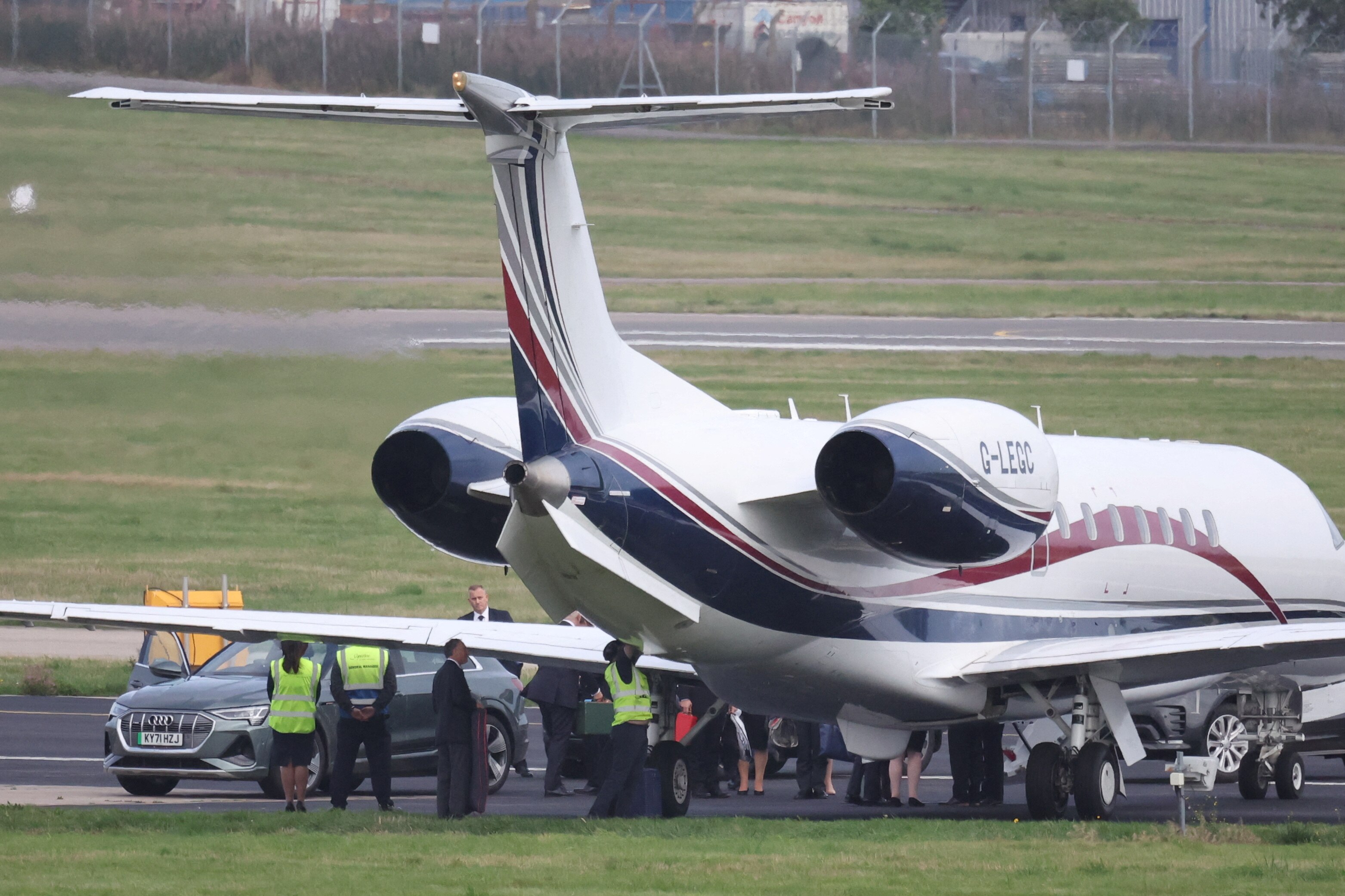 Wide shot of a plane on a tarmac as a silver car pulls up and people crowd beneath it 