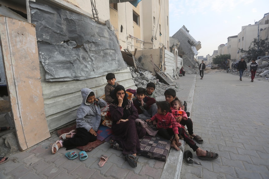 A wide shot of a woman sitting on a footpath in Gaza, sounded by a group of children, near damaged buildings and people walking.