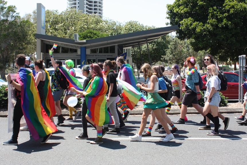 People draping colourful rainbow flags marching down a street with trees in background