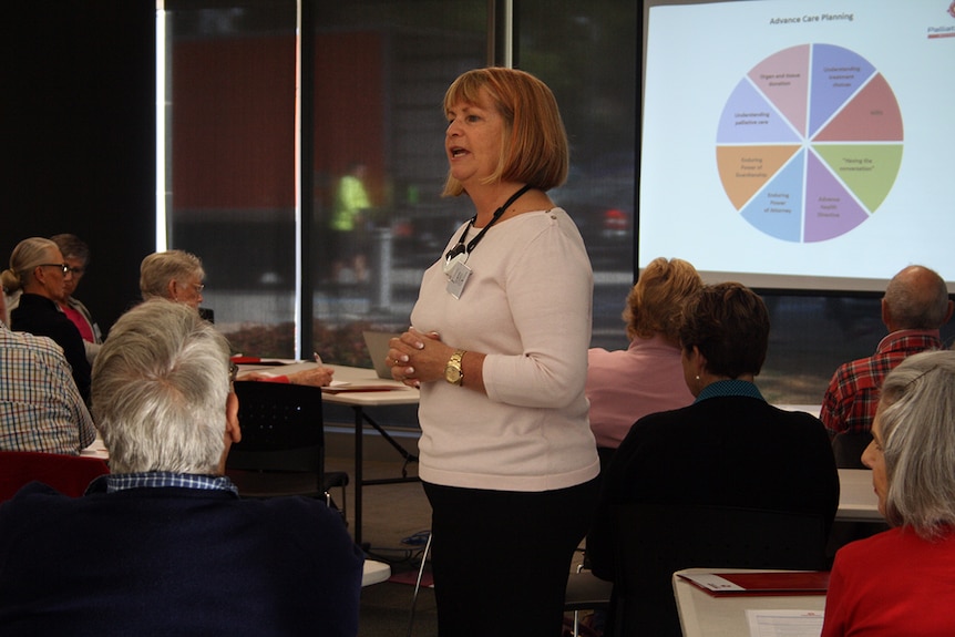 Lana stands in the middle of a room with elderly people, and a projector screen at the front.