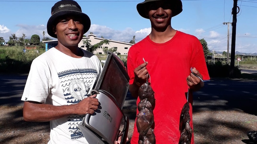 Two men stand next to each other with fish and an electronic item in hand for exchange