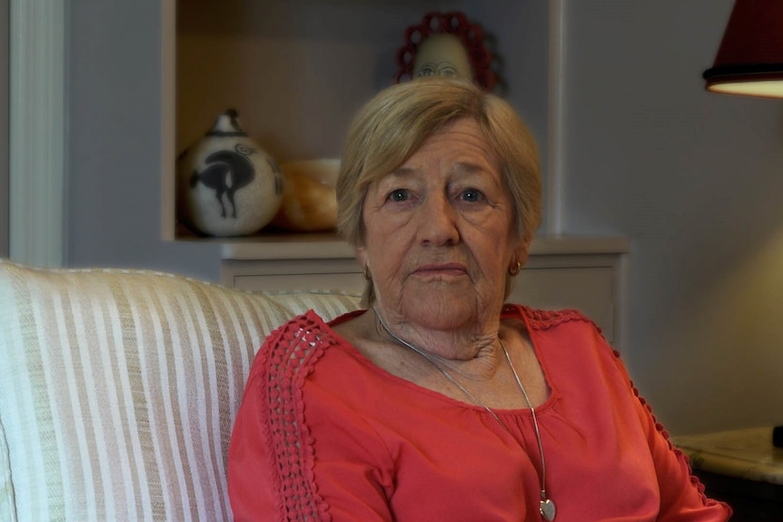 An older woman sits in an armchair, looking solemn.