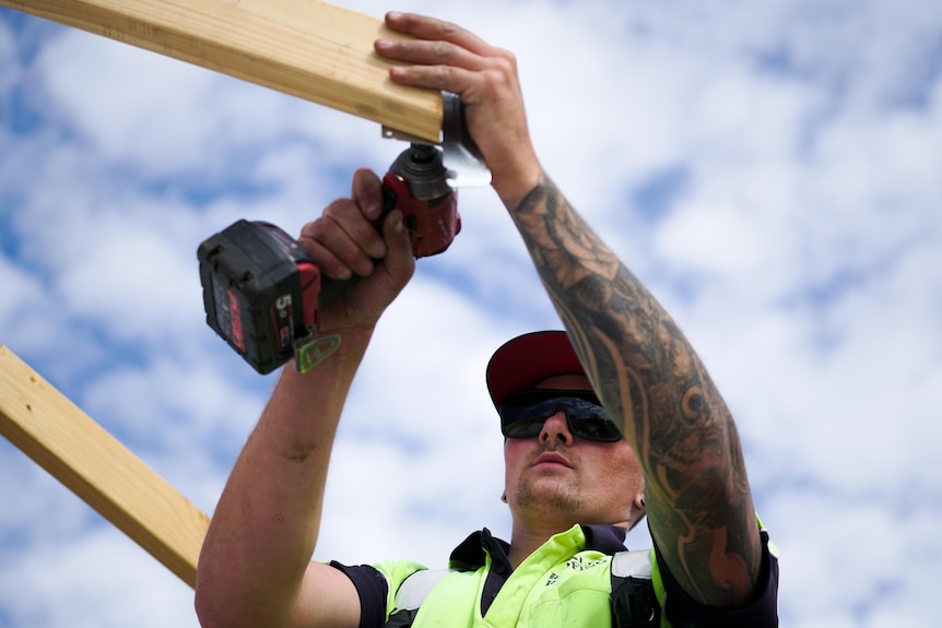A man uses a drill on a wooden frame of a roof