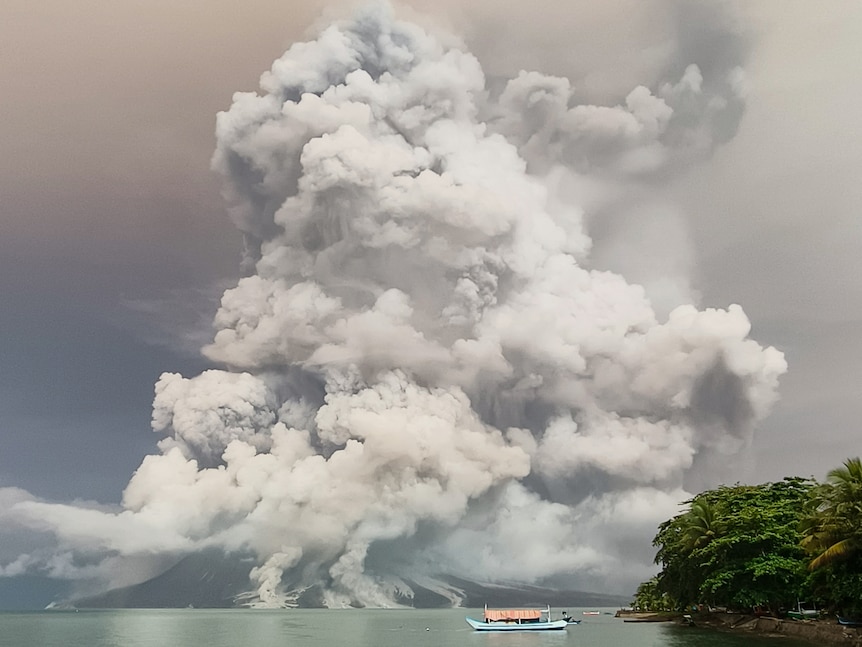 Grey ash spews into the sky above a mountain. A boat sits in the ocean in the foreground.