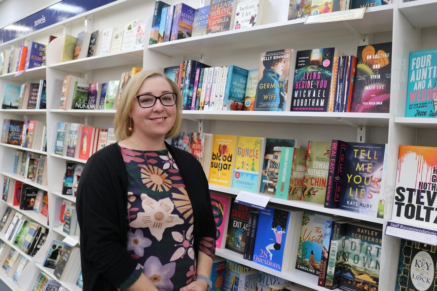 A woman with glasses stands in front of a row of stocked bookshelves.