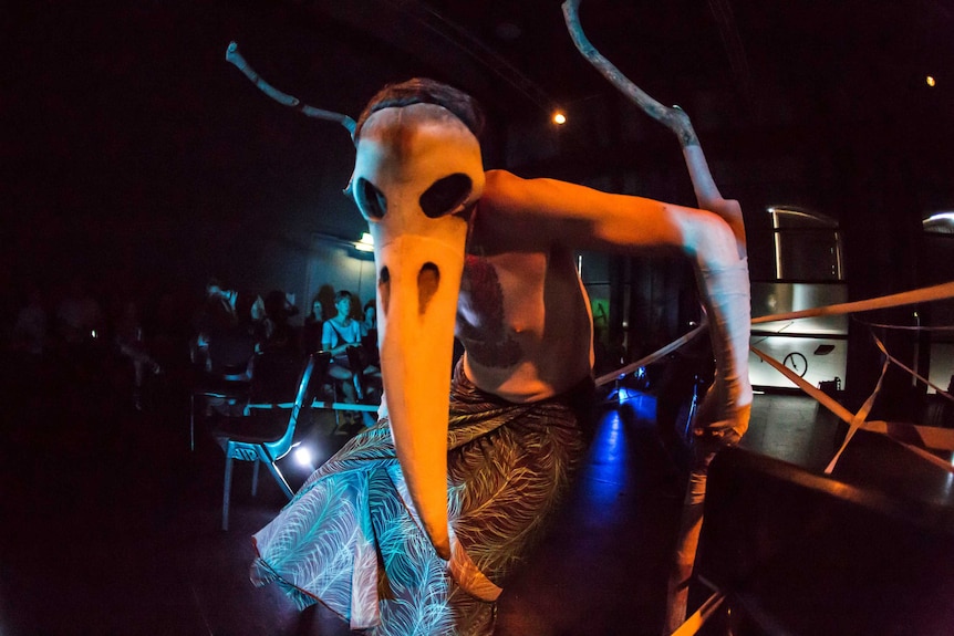 A person in a large skull mask, wearing a skirt, performs in front of an audience.