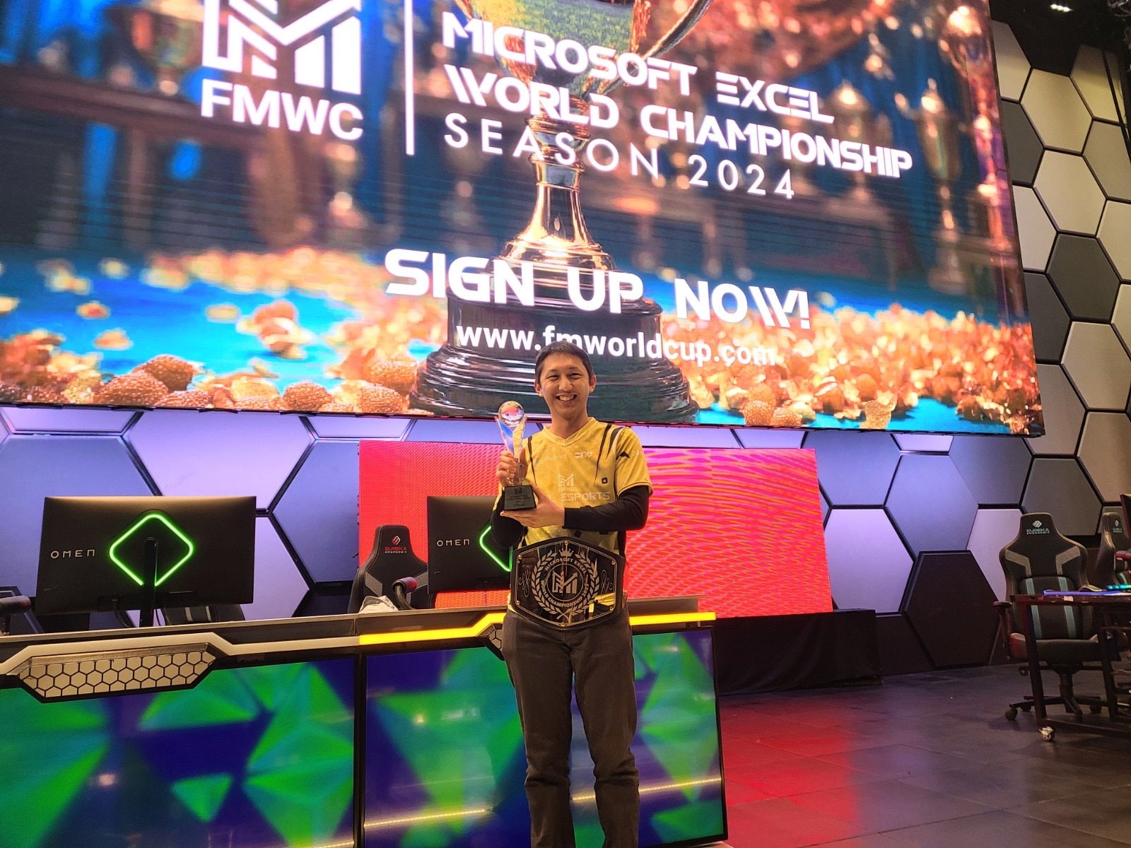 A wide shot of a man of Asian descent holding a trophy in front of two computers and a large screen at an e-sports event