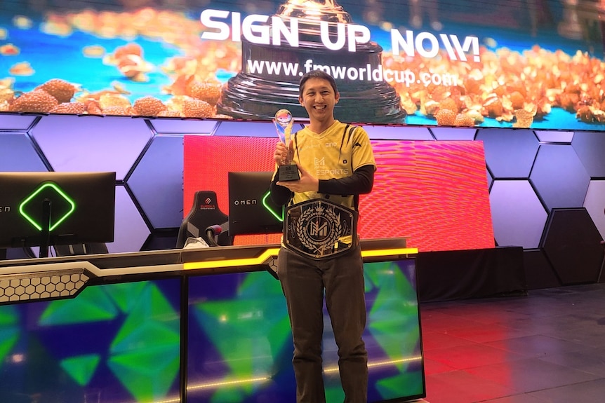 A wide shot of a man of Asian descent holding a trophy in front of two computers and a large screen at an e-sports event