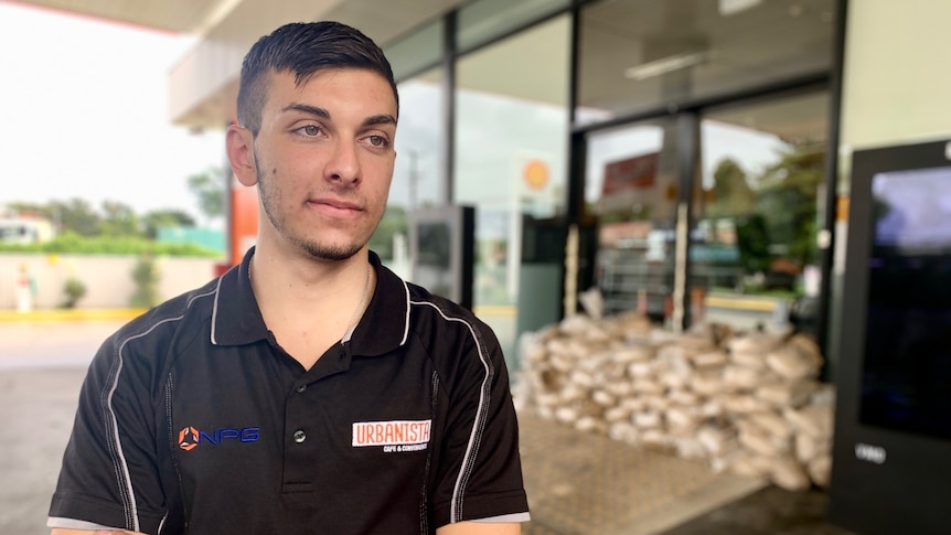 Man stands with arms folded in front of a service station doorway barricaded with sandbags.