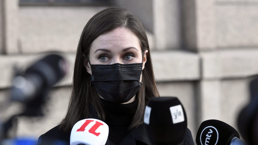 A young female politician wears a black face mask as she speaks into the mics of assembled media.