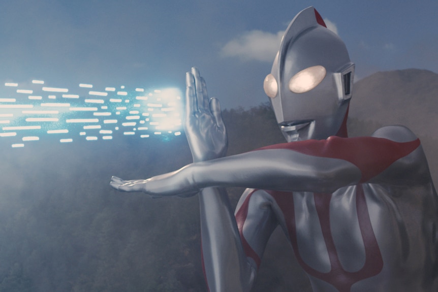 Shin Ultraman, a robot-like superhero in a full-body silver and red suit with helmet, shoots bright blue sparks from his hands.