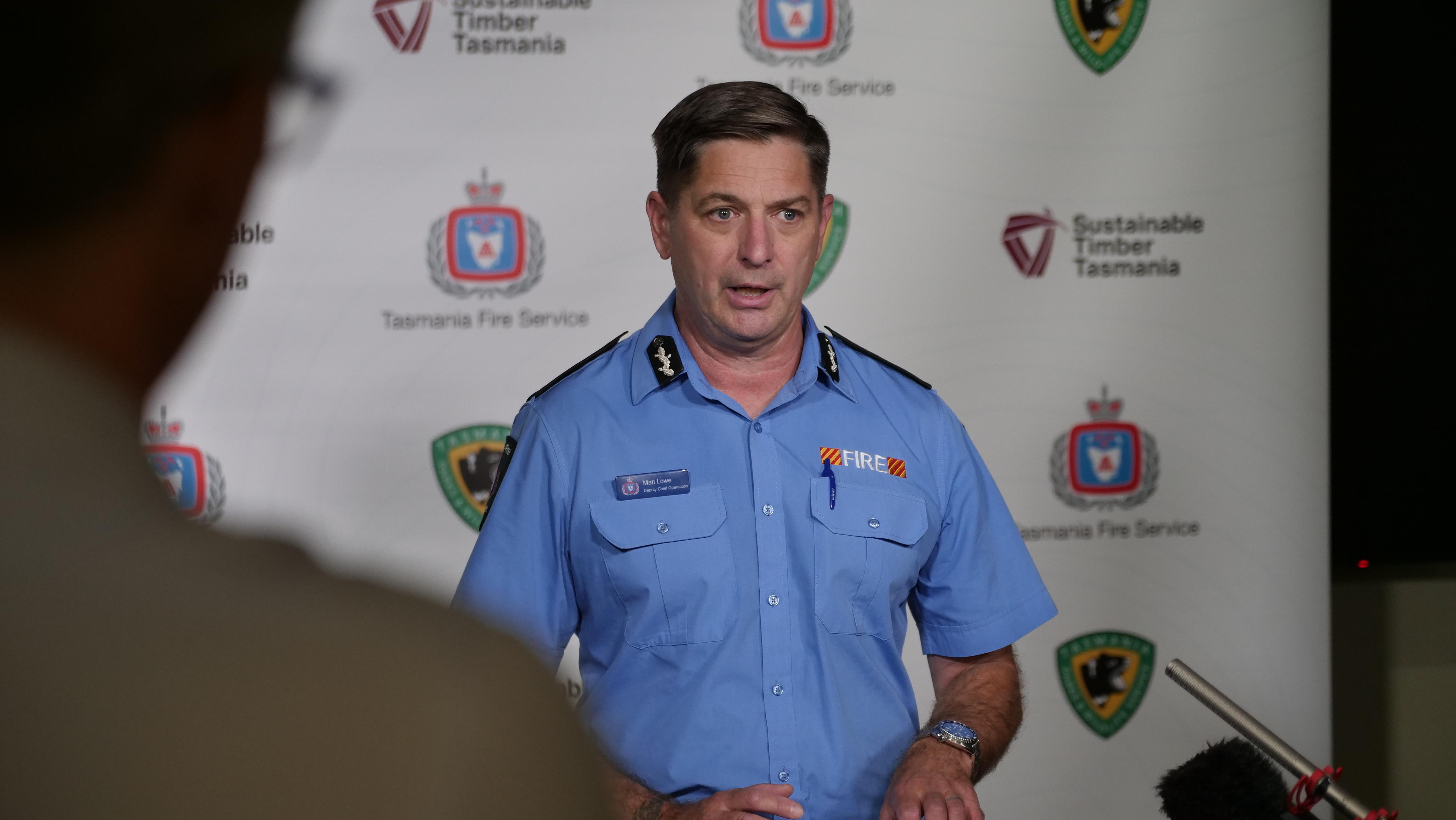 A middle aged man in a blue button up t-shirt gives a press conference. 