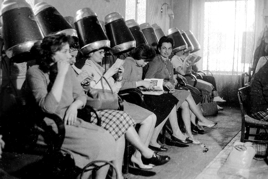 een vintage zwart-wit foto van vrouwen die in een kapsalon zitten en hun haar laten drogen.