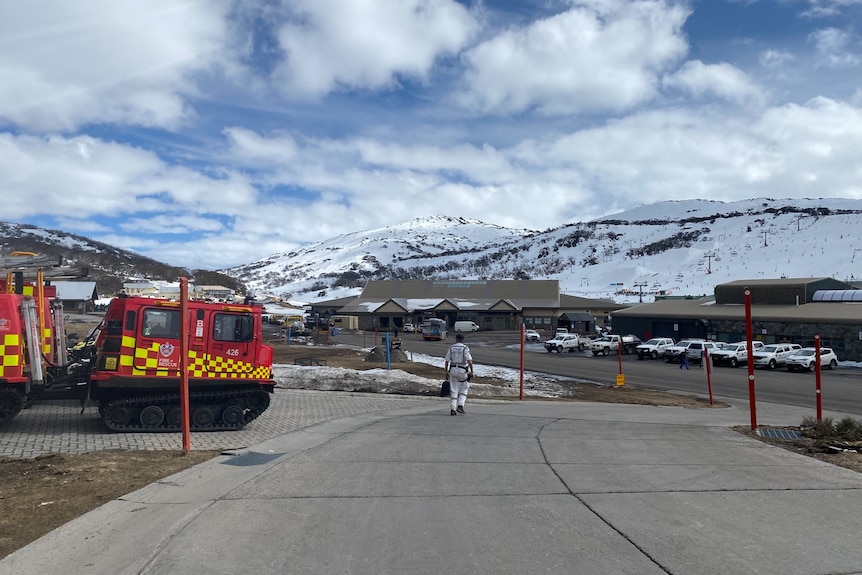The carpark of a ski resort with a fire truck and a police rescuer walking towards snow covered mountains.
