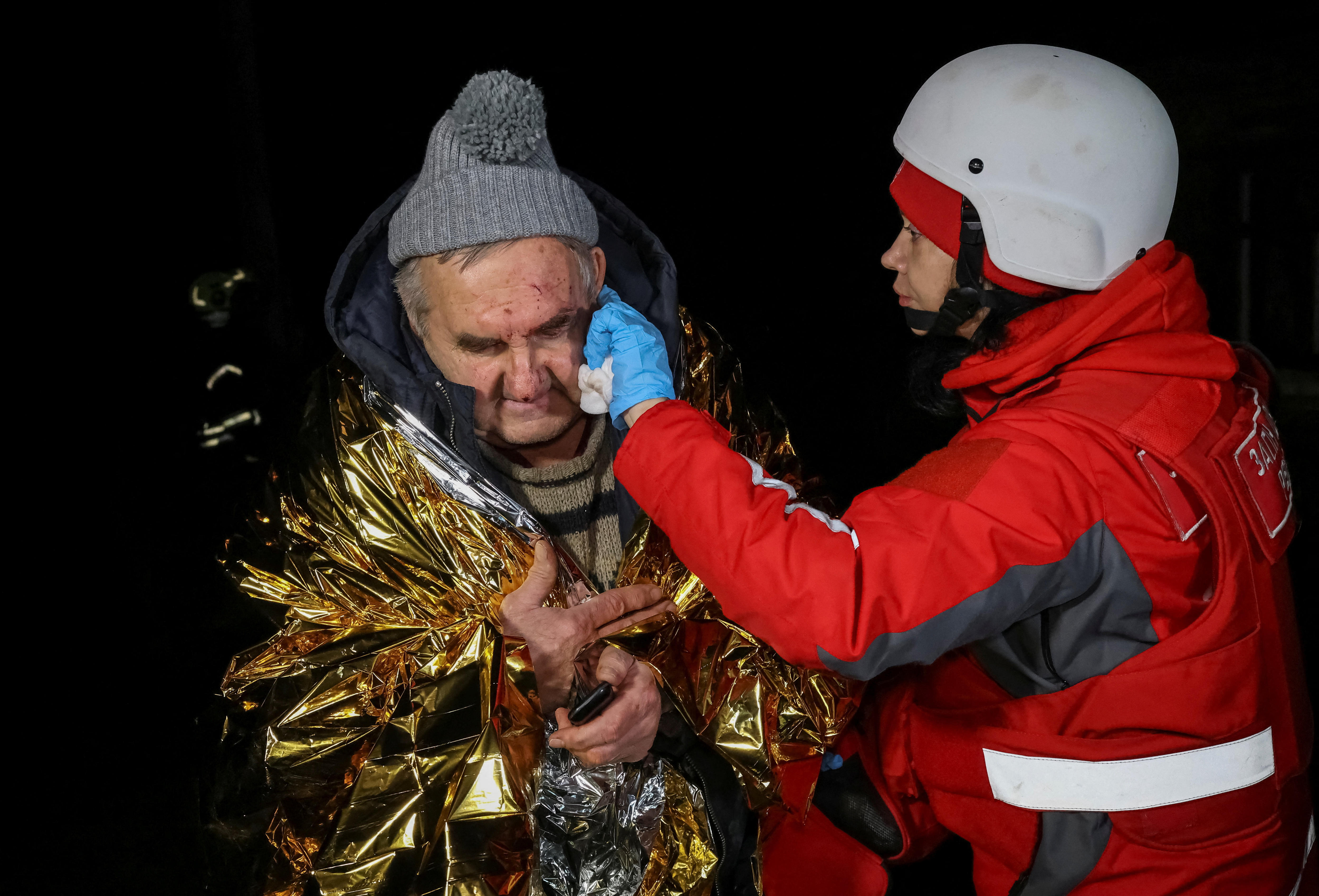 A first responder wipes blood off the face of an older man wrapped in a space blanket.