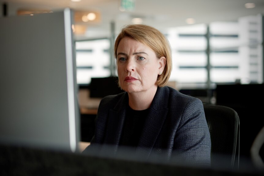 A lady in a dark suit looking worried at her computer desk.