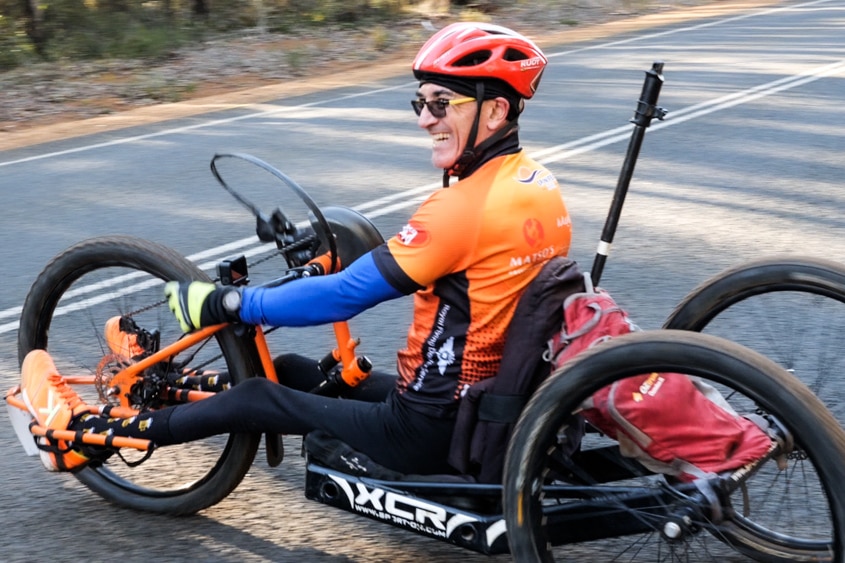 A man in an adaptive, three-wheel mountain bike riding across a road while smiling. 