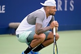Nick Kyrgios crouches down and leans on his racquet on court