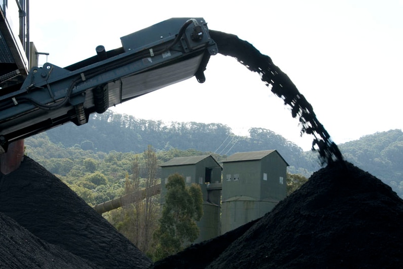 Mining equipment pouring coal onto a heap, wooden buidings in the background, hills, and trees.
