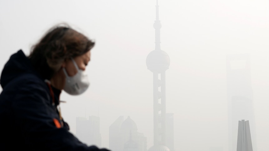Side-on shot of woman wearing a mask and riding a bike with pollution Shanghai in the background.