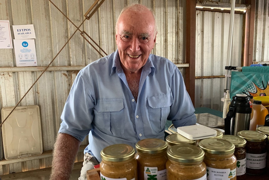 An older man in a blue shirt stands behind a market stall.