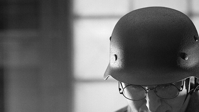 Black and white photographic artwork of a soldier looking down and standing in uniform and hard helmet in dimly lit interior.