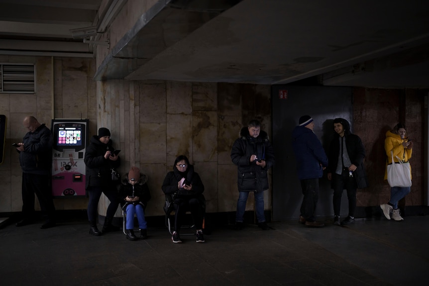 A group of men and women of diverse ages, all wearing winter clothing, gather in a dark concrete tunnel, looking at phones.