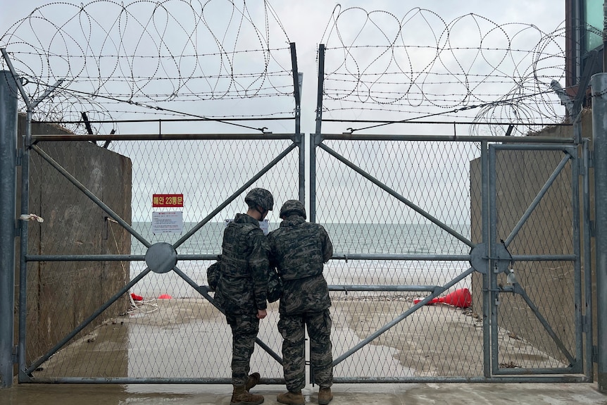 Two men in military uniform standing in front of a fence with barbed wire at the top. 