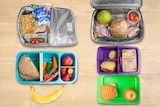 four open lunch boxes with fruit and packets of snacks
