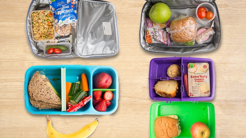 four open lunch boxes with fruit and packets of snacks