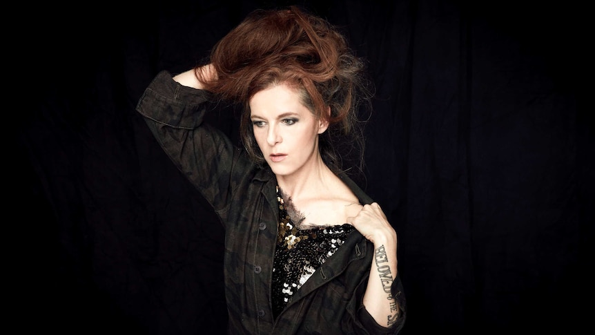 Neko Case stands against a black curtain. One arm is behind her head and the other is pulling her shirt to reveal her shoulder.