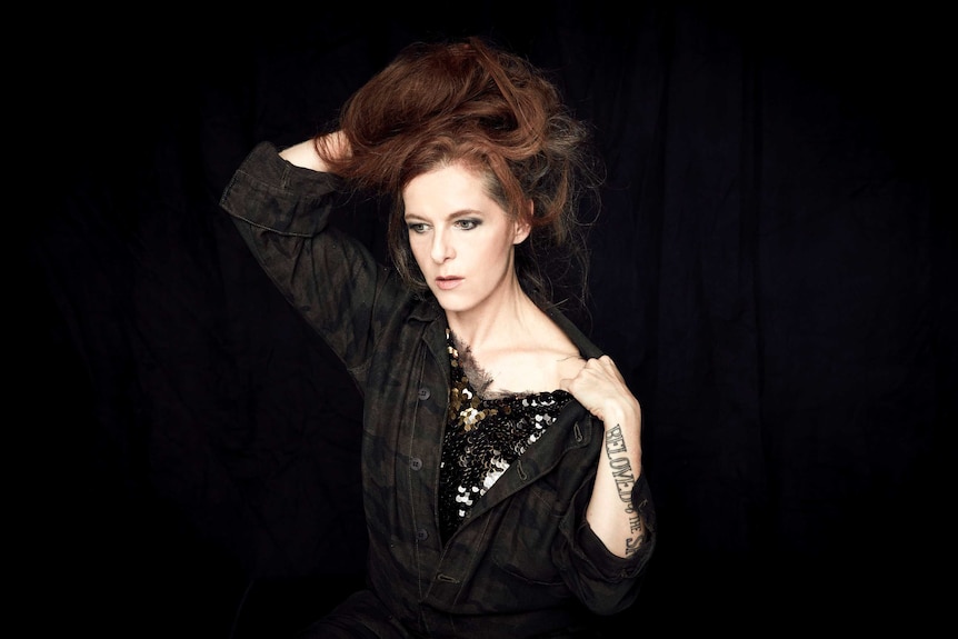 Neko Case stands against a black curtain. One arm is behind her head and the other is pulling her shirt to reveal her shoulder.